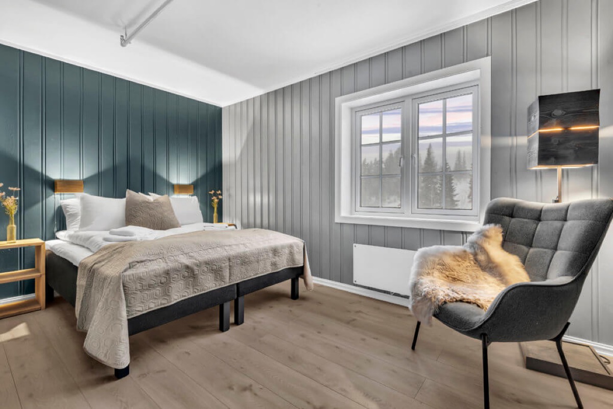 Soverom med grønn panelvegg, saueskinn over lenestol og vindu mot skog