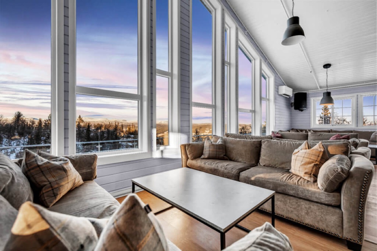 Stor fellesstue med sofagrupper og panoramavinduer mot fjellet i solnedgang